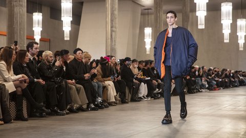 "Ein Model läuft über einen Laufsteg in einem hellen Raum mit hohen Säulen und warmem Licht. Es trägt ein dunkles Outfit mit auffälligem, orangefarbenem Innenfutter. Das Publikum sitzt dicht gedrängt an beiden Seiten und verfolgt die Präsentation."