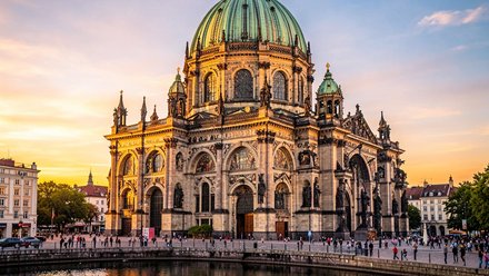 Der Berliner Dom an der Spree bei Sonnenuntergang, große grün patinierte Kuppel und reich verzierte Fassaden, spiegelnde Wasserfläche im Vordergrund und Passanten am Ufer.