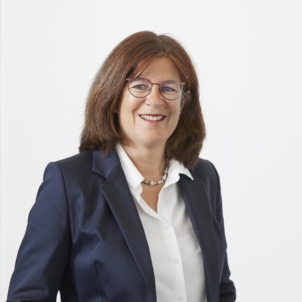 Ein Porträt zeigt eine Person vor hellem, neutralem Hintergrund in formeller Kleidung, mit ruhigem Blick und freundlichem Ausdruck, die klare Bildkomposition lenkt den Fokus auf Präsenz und Professionalität, wodurch das Bild für fachliche Verantwortung, Expertise und die menschliche Perspektive innerhalb von Architektur, Planung und institutioneller Arbeit steht.