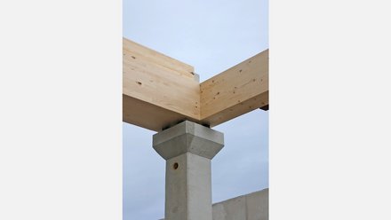 Nahes Anschlussdetail: Zwei helle Brettschichtholzträger treffen über Eck und lagern auf einem geformten Betonstützenkopf vor grauem Himmel.