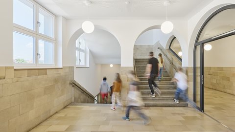 Treppenhaus mit hellen Wänden, großen Fenstern, runden Deckenbögen und mehreren Kindern, die die Treppe hinauf- und hinuntergehen