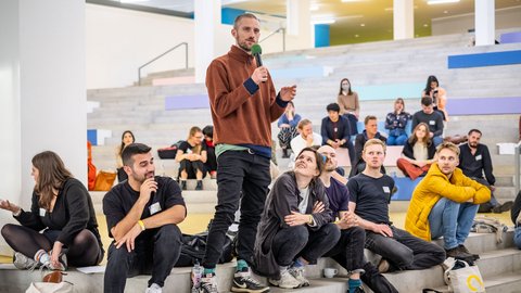 Person spricht mit Mikrofon vor einer Gruppe von Veranstaltungsteilnehmenden.