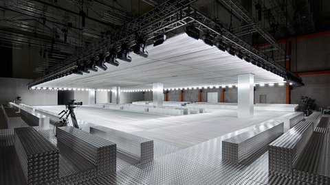 "Moderner Laufsteg aus silbernem Metall mit strukturiertem Boden. Über der Fläche hängt eine große rechteckige Lichtinstallation mit vielen Scheinwerfern. Kameras stehen am Rand bereit für Aufnahmen. Der Raum hat eine industrielle, technische Ästhetik."