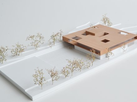 Architekturmodell: Flacher, holzfarbener Baukörper mit quadratischen Öffnungen kragt über weißem Sockel; umlaufend Wege und kleine Modellbäume.