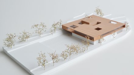 Architekturmodell: Flacher, holzfarbener Baukörper mit quadratischen Öffnungen kragt über weißem Sockel; umlaufend Wege und kleine Modellbäume.