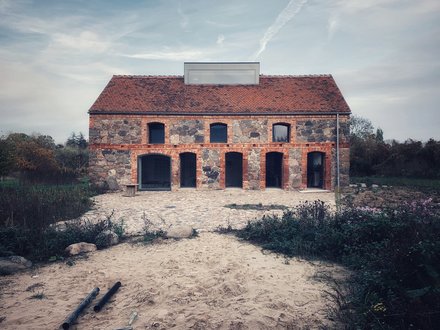 Frontansicht Feldsteinhaus Uckermark: Zweigeschossiger Bau mit rotem Ziegeldach, Arkaden im Erdgeschoss; Pflasterhof und sandiger Vorplatz.