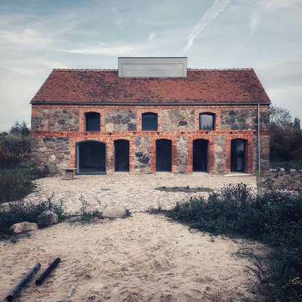 Frontansicht Feldsteinhaus Uckermark: Zweigeschossiger Bau mit rotem Ziegeldach, Arkaden im Erdgeschoss; Pflasterhof und sandiger Vorplatz.