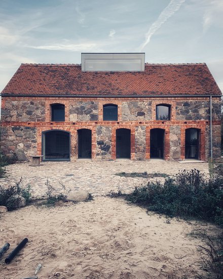 Frontansicht Feldsteinhaus Uckermark: Zweigeschossiger Bau mit rotem Ziegeldach, Arkaden im Erdgeschoss; Pflasterhof und sandiger Vorplatz.