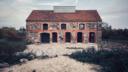 Frontansicht Feldsteinhaus Uckermark: Zweigeschossiger Bau mit rotem Ziegeldach, Arkaden im Erdgeschoss; Pflasterhof und sandiger Vorplatz.