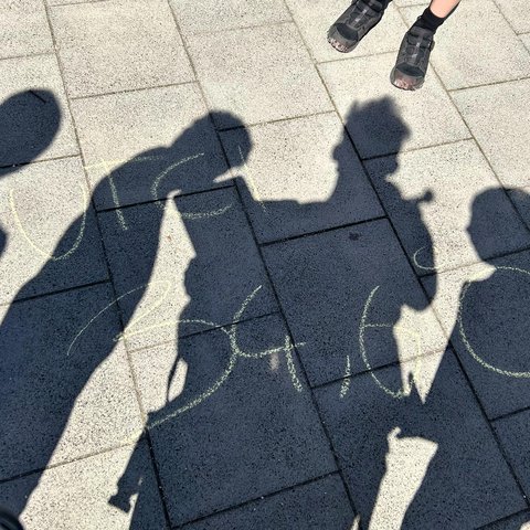 Die Beine mehrerer Personen und der Schattenwurf einer Gruppe auf Steinplatten, auf denen eine Kreideaufschrift zu sehen ist.