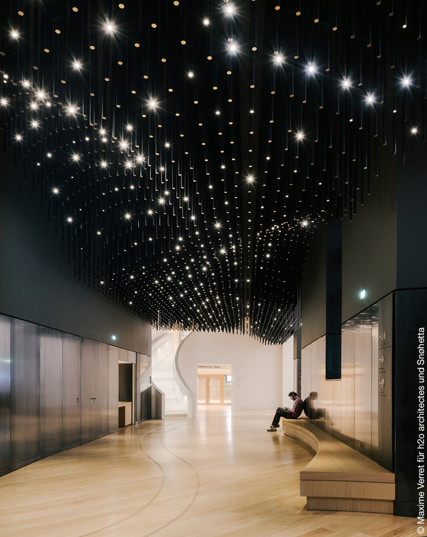 Eingangsbereich eines Museums mit Holzboden, geschwungener Treppe und Lichtstrahlern an der Decke.