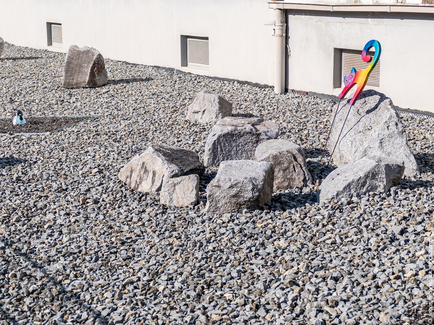 Ein Schottergarten voller Kies, in der eine bunte Figur in Katzenform steht.