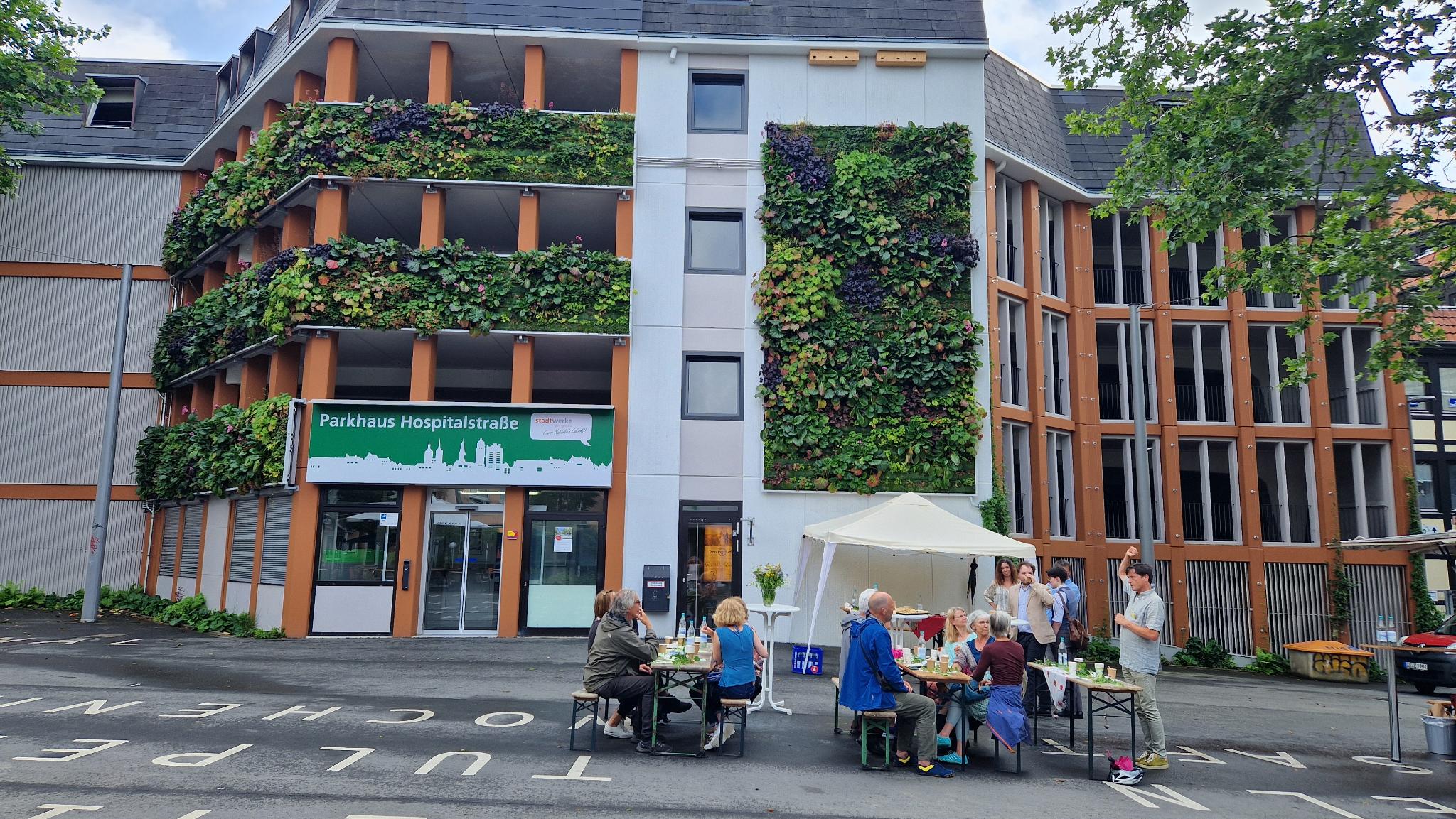 Männer und Frauen sitzen vor einem Haus mit begrünter FAssade.