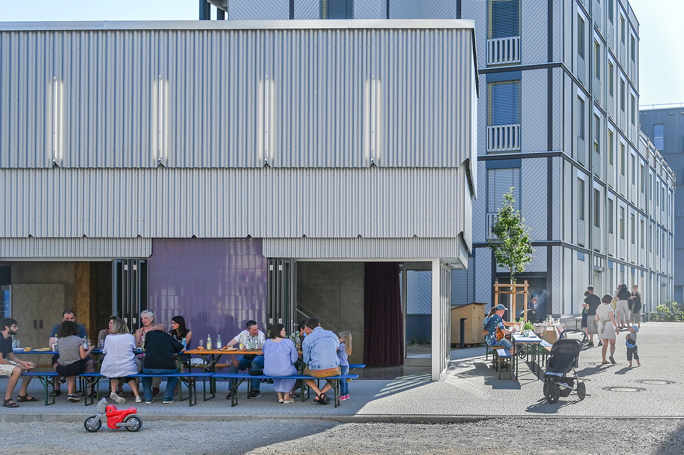 Pavillon mit Fassade aus grauem Wellblech, davor Menschen