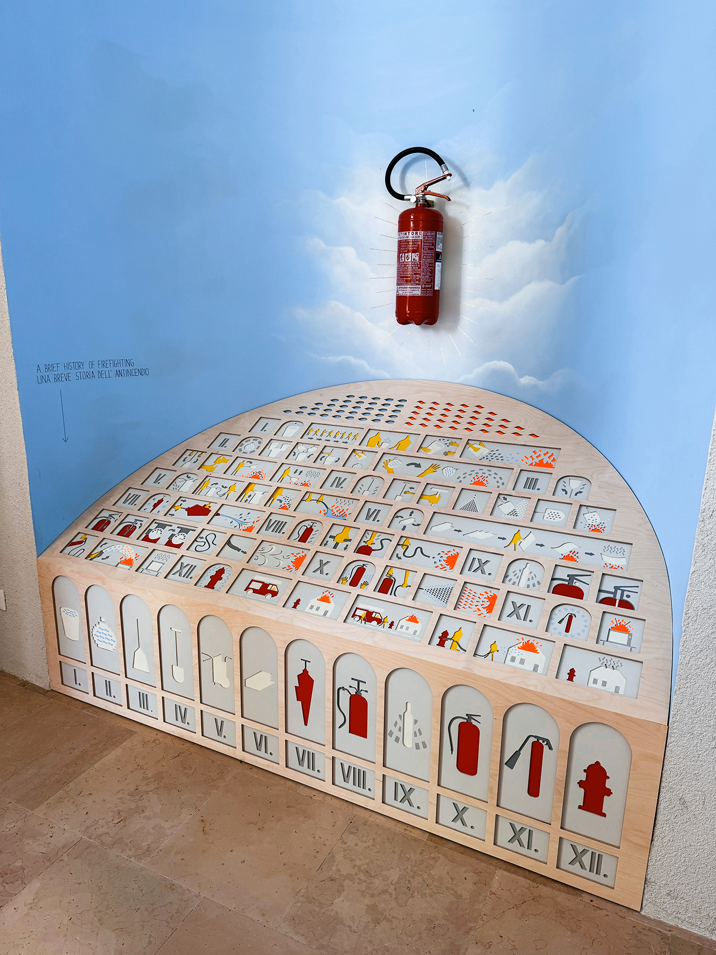 Feuerlöscher vor einer hellblauen Wand, darunter ein Halbrund aus Holz mit Bildern von Feuerlöschern und Notausgängen.