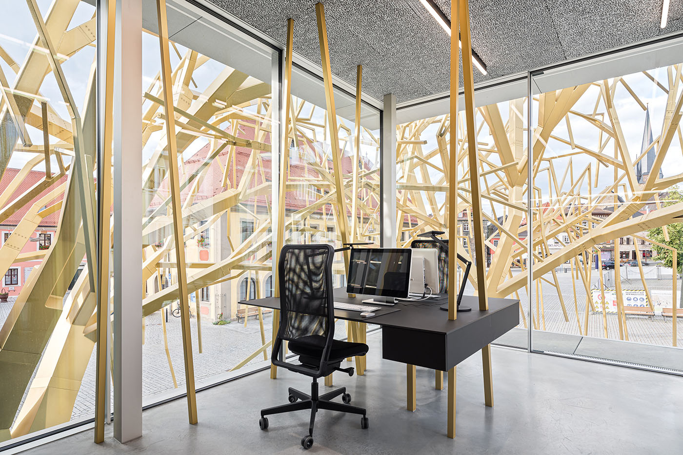 Büro mit bodentiefen Fenstern, einem schwarzen Schreibtisch und schwarzen Schreibtischsesseln und einer Konstruktion aus Holzästen vor dem Fenstern.