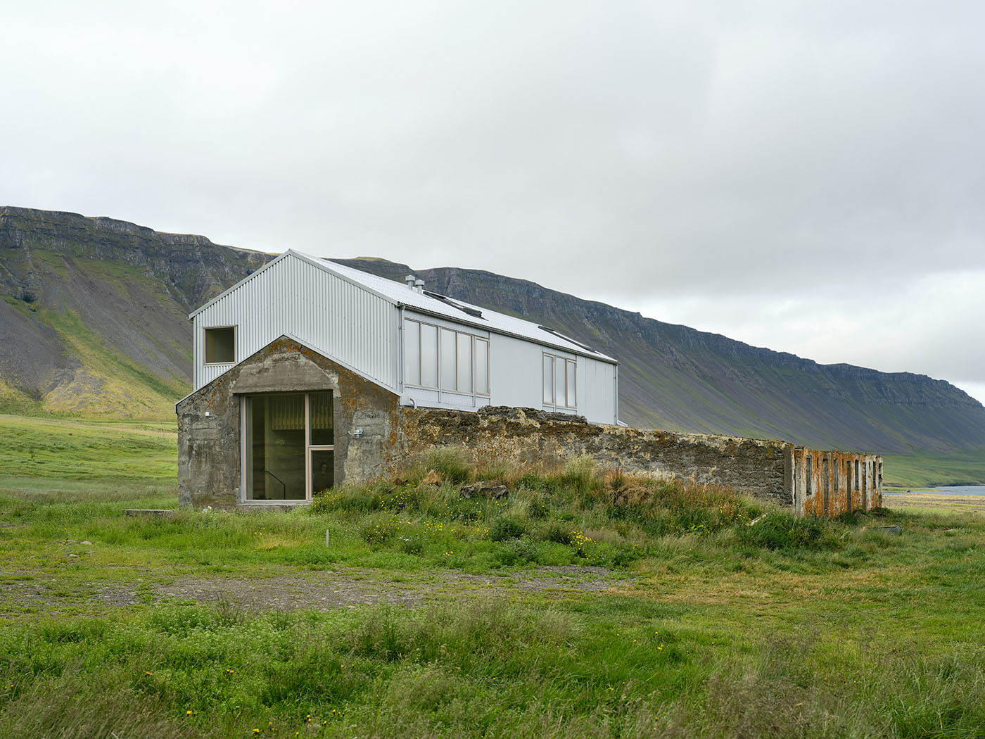 Das Foto zeigt ein Haus in Island auf einer Wiese vor einem Wall aus grau-grünem Gestein